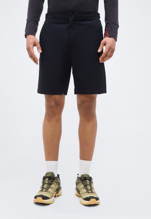 Homme portant un short noir, des chaussettes blanches et des chaussures de sport vertes, debout devant un fond clair uni.