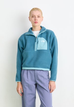 Blauwe fleece pullover met een kwart ritssluiting, voorzien van een lichtblauwe borstzak en manchetten. Model draagt lavendelkleurige broeken.