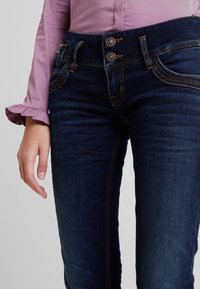 Mörkblå denimjeans med hög midja, med två metallknappar och kontrasterande gul söm längs framseam.