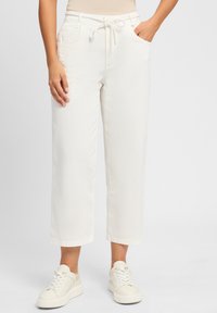 Pantalons larges beige clair, coupe courte, avec une taille à cordon, dotés de poches latérales et d'une texture lisse, associés à des baskets blanches.