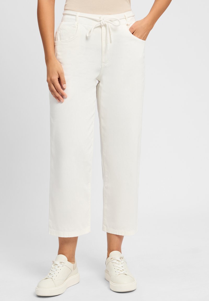 Pantalons larges beige clair, coupe courte, avec une taille à cordon, dotés de poches latérales et d'une texture lisse, associés à des baskets blanches.