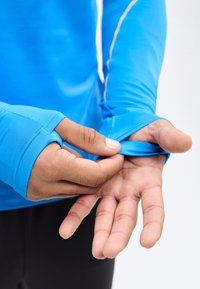 Personne ajustant l'orifice pour le pouce sur la manche d'un haut de sport bleu vif, révélant la paume et les doigts sur un fond uni.