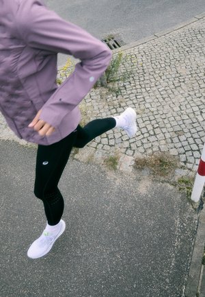 Lilla spordijope koos tekstureeritud disainiga, mustad legginsid ventilatsiooni detailidega, valged jooksujalatsid ja valged sokid liikumises asfalttee peal.