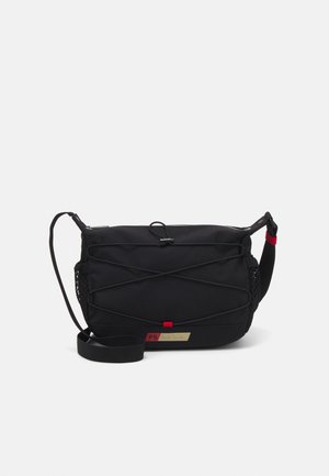 Sac bandoulière noir avec sangle réglable, cordon élastique croisé à l'avant, poches latérales en mesh et petite étiquette de marque rouge et or.