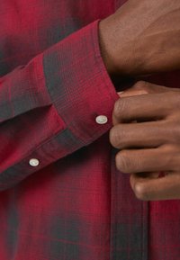 Camisa a cuadros en rojo y negro con mangas largas, que cuenta con puños abotonados y botones blancos, elaborada con tejido texturizado.