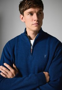 Blauer Zip-Hals-Pullover aus einem weichen, strukturierten Strick. Mit einem dunkelblauen Logo auf der Brust und geribbelten Bündchen, vor einem neutralen Hintergrund.