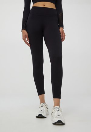 Leggings neri a coste con una vita alta, forma aderente e tessuto texturizzato. Abbinati a scarpe da ginnastica bianche. Sfondo neutro.