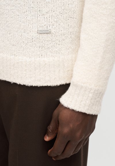 Cream textured sweater featuring ribbed cuffs and hem; small metallic brand tag on the front; worn over dark brown pants.