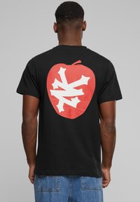 Sort bomulds t-shirt med et stort rødt æblegrafik og et hvidt snefnugdesign på ryggen. Korte ærmer, afslappet pasform.