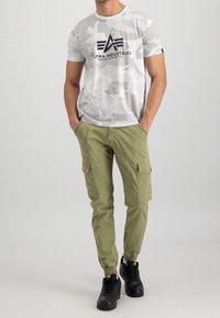 Camiseta blanca de camuflaje con logo negro, combinada con pantalones cargo verde oliva que tienen bolsillos laterales y tobillos ajustados, llevados con zapatillas negras.