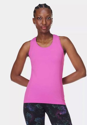 Woman with braided hair wearing a sleeveless pink top and black leggings with a colorful abstract pattern, standing with hands behind her back.