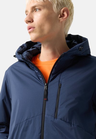 Young person with short blond hair wearing a navy blue hooded jacket over an orange shirt, looking to the left.
