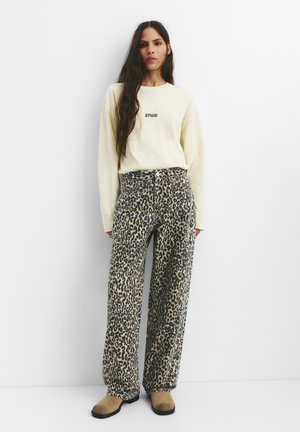 Sweatshirt oversized de culoare galben deschis cu logo-ul "STWD", purtat împreună cu pantaloni largi cu imprimeu de leopard și pantofi slip-on maro.