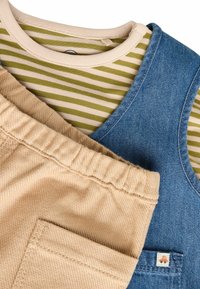 Modré denimo overaly, vrstvené na pruhovanej olivovej a smotanovej košeli. Hnedé kraťasy s textúrovanou tkaninou a malým akcentovým vreckom.