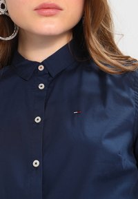 Žena v tmavě modré košili na knoflíky s malým vyšívaným logem v červené, bílé a námořnické modři na hrudi a velkým stříbrným kruhovým náušnicí.