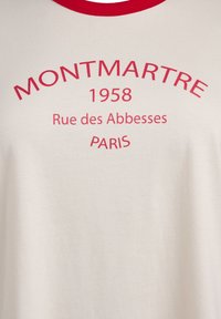 Kremo spalvos trumpų rankovių marškinėliai su raudonu apykaklės kraštu. Priekyje yra raudonas tekstas, skaitantis "MONTMARTRE 1958 Rue des Abbesses PARIŽIUS."