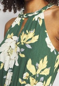 Kvinna klädd i en grön halterneck-klänning med stora vita och gula blommor, flätad axelrem och plisserad tyg.