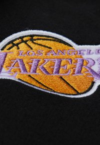 Schwarzer Stoff mit einem gestickten Logo und einem Basketball-Graphic in Orange, Text "LOS ANGELES LAKERS" in Lila, eingerahmt von weißem Nähen.