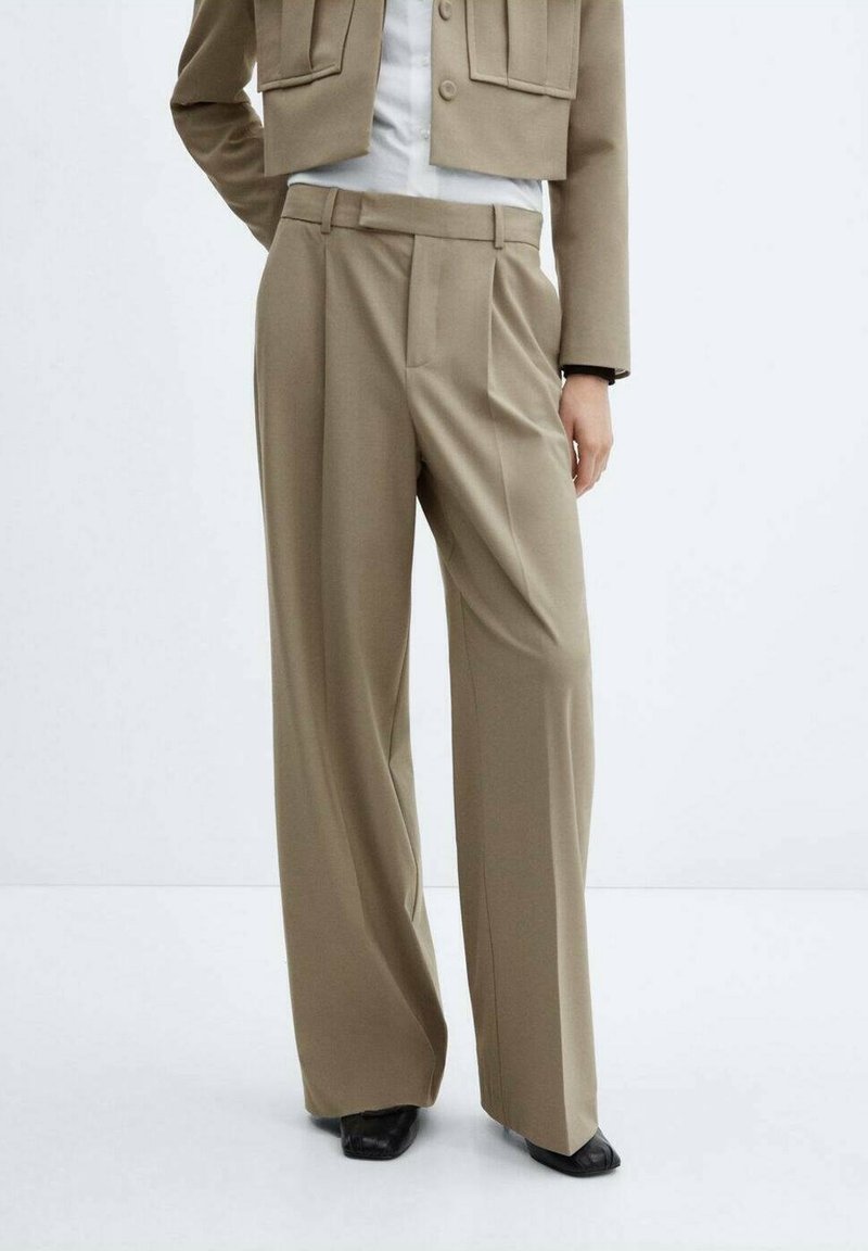 Pantalon beige taille haute à jambes larges associé à des chaussures noires à bout carré et une veste courte beige portée sur une chemise blanche.