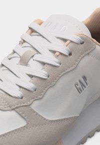 Baskets blanches et beiges avec lacets blancs, accents en daim et logo "GAP" embossé sur le côté.