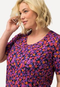 Tričko s krátkým rukávem vyrobené z látky s černým podkladem a živým květinovým vzorem v purpurových, růžových, oranžových a žlutých tóninách. Výstřih ve tvaru lodi.