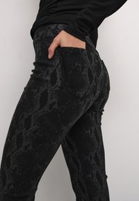 Zwarte slangenprint leggings met een gladde textuur, voorzien van achterzakken en een getailleerde pasvorm. Het materiaal lijkt rekbaar en figuurvormend.