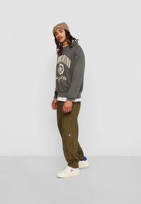 Felpa grigia con scritte dorate, pantaloni verde oliva con cerniere e sneakers bianche con accenti blu. Un berretto beige completa l'outfit.