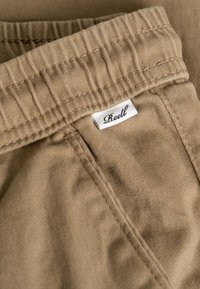 Primer plano de la cintura de unos pantalones beige con dobladillo elástico y una pequeña etiqueta de tela blanca con la marca "Roell" cerca del bolsillo cosido.