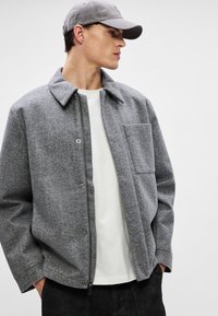 Grau wollene Jacke mit Kragen, Reißverschluss vorne und einer Brusttasche. Kombiniert mit einem weißen Hemd und dunklen Jeans, unter einer grauen Mütze.