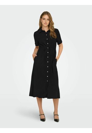 Nainen mustassa napillallisessa midi-mekossa, jossa on lyhyet hihat, vyö vyötäröllä, kädet taskuissa ja mustat Mary Jane -korkokengät.