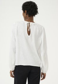 Blouse blanche avec une texture à motifs, manches longues, un dos en forme de clé avec un lien de fermeture, et une coupe ample, assortie à un pantalon noir.