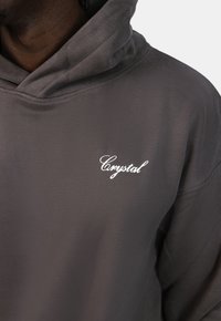 Šedá mikina s měkkou texturou, šňůrkovou kapucí a malým bílým výšivkovým logem "Crystal" na hrudi.