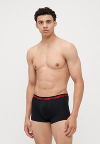 Boxer shorts en coton noirs avec une ceinture à logo HUGO rouge. Présentent une coupe ajustée et une texture lisse. Le modèle se tient contre un fond neutre.
