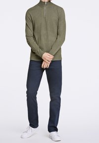 Sweat à zip olive avec manches longues, bord côtelé, associé à un pantalon bleu marine et des baskets blanches, se tenant devant un fond neutre.