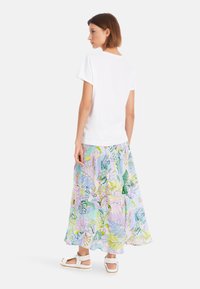 Weißes Kurzarm-T-Shirt, kombiniert mit einem bunten, gemusterten langen Rock in Pastellfarben und Illustrationen. Leichte Sandalen runden den Look ab.