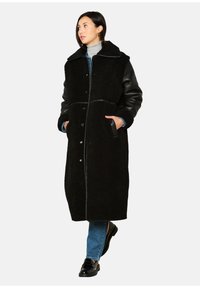 Zwarte lange jas met een schapenvachtstructuur, leren mouwen, kraag en knoopsluiting aan de voorkant. Model draagt een spijkerbroek en glanzende zwarte schoenen.