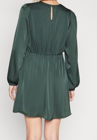 Vihreä satiini mekko pitkillä hihoilla, kootulla vyötäröllä ja avonaisella selkäyksityiskohdalla. Korkealaatuinen pinta ja virtaava siluetti.
