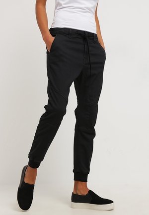 Person wearing black jogger pants with side pockets and black slip-on shoes, standing against a plain light background.