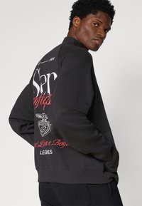 Black sweatshirt featuring graphic print on the back in red and white, ribbed cuffs, and a relaxed fit. Soft texture, casual design.