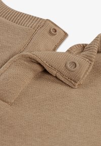 Vaaleanruskea neulepaita, jossa on resorikaulus. Mallissa on olkapäillä napilliset korostukset ja pehmeä tekstuuri. Kankaan yksityiskohtainen ompelu on nähtävissä.