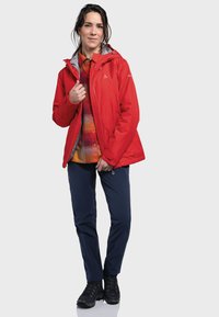 Red waterproof jacket with gray lining, featuring a hood and front zipper. Worn over a plaid shirt, paired with navy pants and black boots.
