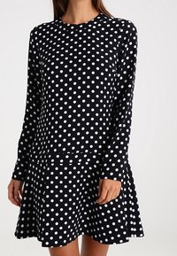 Femme portant une robe noire à manches longues avec des pois blancs, dotée d'un col rond et d'une jupe évasée.