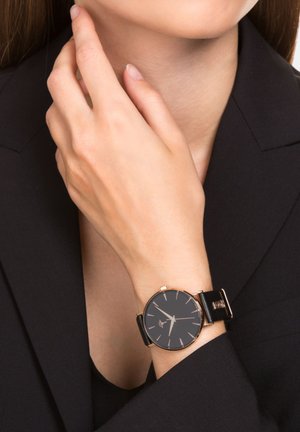 Handgelenk mit einer schwarzen Lederuhr, die ein schwarzes Zifferblatt und goldene Akzente aufweist, getragen von einer Person in einem schwarzen Blazer, die sich am Hals berührt.