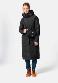 Zwarte lange jas met capuchon, voorzien van een ritssluiting aan de voorkant en zakken. Gecombineerd met oranje laarzen en een kleine zwarte crossbodytas.