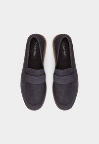 Paire de mocassins en daim noir avec semelles tissées couleur fauve, semelles intérieures noires estampillées Calvin Klein, vues de dessus sur un fond blanc.