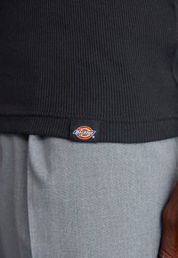 Juodas raštuotas jersey marškinėlis su matomu "Dickies" logotipu apačioje, derinamas su šviesiai pilkais audinio kelnais. Dėmesys tekstūrai ir spalvų kontrastui.