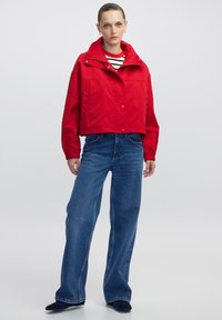 Vrouw die staat in een rode cropped jas over een zwart-wit gestreept shirt, met wijde blauwe jeans en zwarte schoenen op een effen achtergrond.