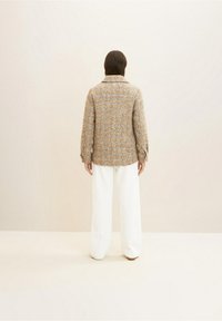 Een persoon met schouderlang bruin haar, gekleed in een tan en grijs geruite jas en witte wijde broek, staand tegen een beige achtergrond.