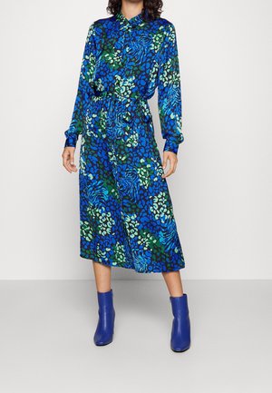 Kvinde iført en langærmet midikjole med blå, grønne og sorte mønstre samt bindebånd i taljen, kombineret med blå ankelstøvler.