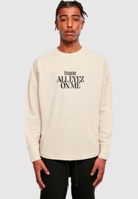 Sweatshirt bej cu mânecă lungă și text negru care spune "tupac ALLEYEZ ON ME", având un guler tip crew și o croială relaxată. Pantaloni negri vizibili.
